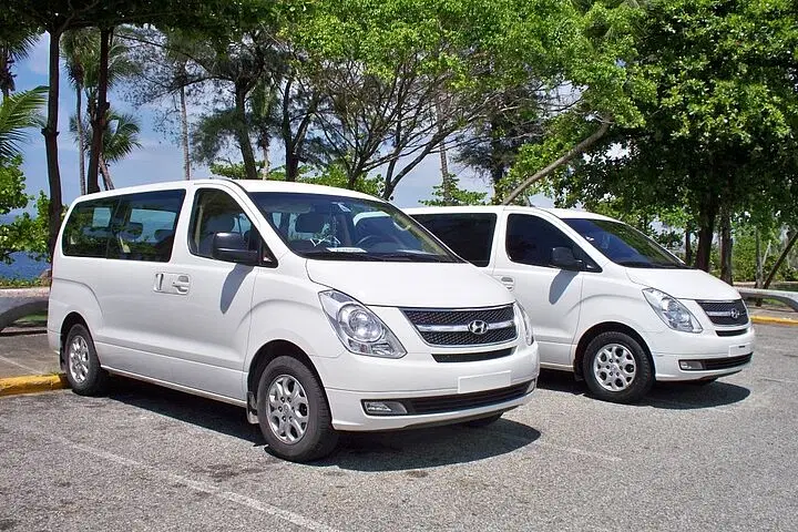 Two white Hyundai vans, perfect for tours in Punta Cana, are parked side by side in a sunny outdoor lot. Green trees, blue water, and a clear sky set the scene. Both vans have tinted windows and face right, ready for your next Punta Cana trip.