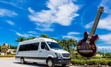 A white passenger van is parked on a sunny road beside the iconic Hard Rock Hotel & Casino guitar sign—perfect for starting unforgettable punta cana excursions. Palm trees and blue skies set the tropical, welcoming scene.