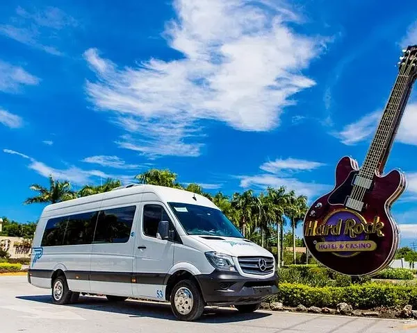A white passenger van is parked on a sunny road beside the iconic Hard Rock Hotel & Casino guitar sign—perfect for starting unforgettable punta cana excursions. Palm trees and blue skies set the tropical, welcoming scene.
