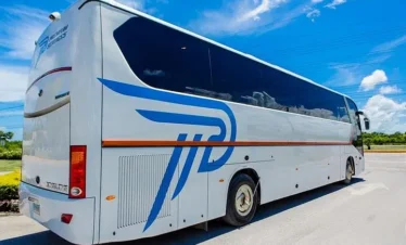 A modern white tour bus with large tinted windows and a blue and orange logo is parked on a paved road under a bright blue sky—perfect for unforgettable Punta Cana tours. Green grass and utility wires can be seen in the background.