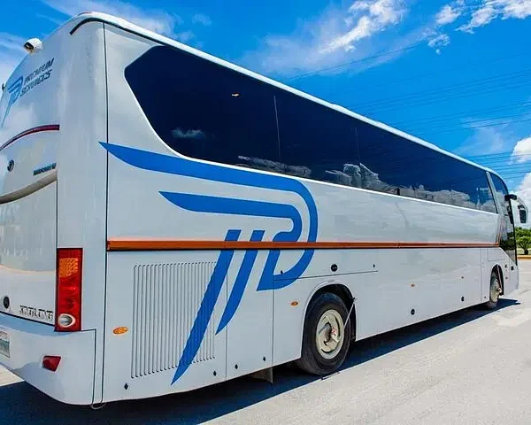 A modern white tour bus with large tinted windows and a blue and orange logo is parked on a paved road under a bright blue sky—perfect for unforgettable Punta Cana tours. Green grass and utility wires can be seen in the background.