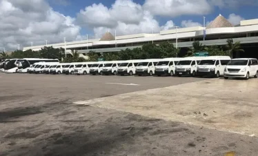 A row of white passenger vans is parked side by side in a large parking lot under a partly cloudy sky, ready for Punta Cana tours. Behind the vans, a modern building and lush greenery hint at an airport or transport hub setting.
