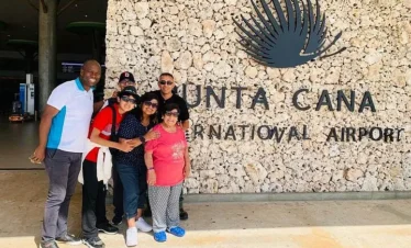 Five people stand smiling in front of a stone wall displaying "Punta Cana International Airport." Ready for their Punta Cana trip, the group appears happy, posing together in the sun with dreams of tours en Punta Cana ahead.