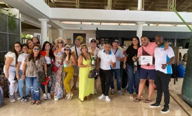 A group of about 20 adults pose indoors at a resort lobby with marble floors, smiling at the camera. Most wear casual summer clothes and sunglasses, some holding drinks—a fun start to one of their Punta Cana excursions. One man holds a “P Quintero” sign.