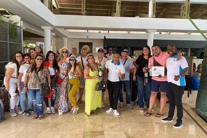 A group of about 20 adults pose indoors at a resort lobby with marble floors, smiling at the camera. Most wear casual summer clothes and sunglasses, some holding drinks—a fun start to one of their Punta Cana excursions. One man holds a “P Quintero” sign.