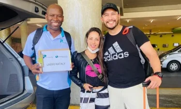 Three people stand smiling outside near parked vehicles. The man on the left holds a "Lasso Nataly" sign. They could be ready for one of the exciting tours in Punta Cana, perfect for any Punta Cana trip adventure.