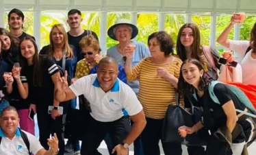A diverse group of people, of various ages and genders, smile and pose together indoors in front of large windows with greenery outside. Some are holding drinks or backpacks, celebrating the start of their Punta Cana tours in a joyful atmosphere.