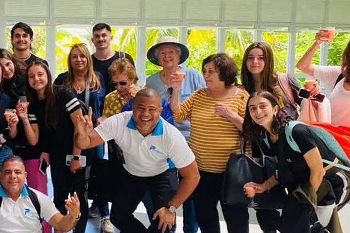 A diverse group of people, of various ages and genders, smile and pose together indoors in front of large windows with greenery outside. Some are holding drinks or backpacks, celebrating the start of their Punta Cana tours in a joyful atmosphere.