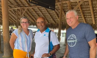Three smiling adults stand under a thatched roof at an airport arrival area, ready for punta cana tours. The man in the center holds a “Marta/Taveri” sign, while signs above point to the exit, promising exciting Punta Cana excursions ahead.