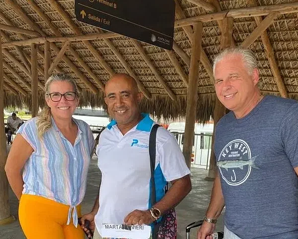 Three smiling adults stand under a thatched roof at an airport arrival area, ready for punta cana tours. The man in the center holds a “Marta/Taveri” sign, while signs above point to the exit, promising exciting Punta Cana excursions ahead.