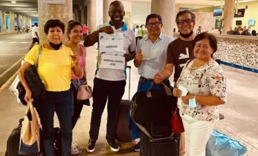 Six smiling adults stand together at an airport arrivals area with luggage. The man in the center holds a sign reading "Andrade Narvaez Maria Jose." Happy and welcoming, they appear ready to begin their unforgettable punta cana tours adventure.