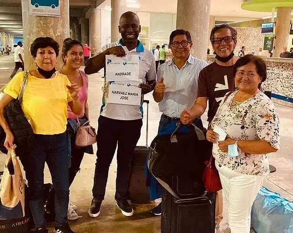 Six smiling adults stand together at an airport arrivals area with luggage. The man in the center holds a sign reading "Andrade Narvaez Maria Jose." Happy and welcoming, they appear ready to begin their unforgettable punta cana tours adventure.