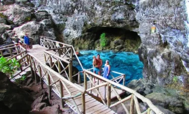A wooden walkway with handrails winds through rocky terrain beside a clear blue natural pool. Two people stand smiling on the walkway near the water—an idyllic moment from their Punta Cana trip, surrounded by jagged rock walls, a cave, and lush greenery.