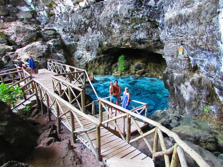 A wooden walkway with handrails winds through rocky terrain beside a clear blue natural pool. Two people stand smiling on the walkway near the water—an idyllic moment from their Punta Cana trip, surrounded by jagged rock walls, a cave, and lush greenery.
