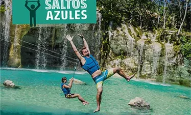 Two people zipline over turquoise water in front of a waterfall on one of the thrilling Punta Cana excursions. Both wear life jackets and swimwear; one has arms raised in excitement. A "SALTOS AZULES" sign sits above, with lush trees surrounding the falls.
