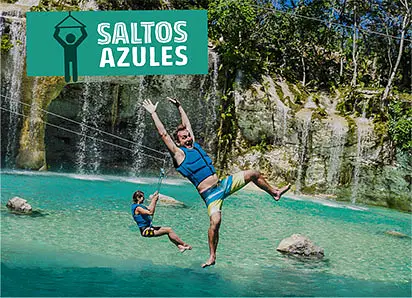 Two people zipline over turquoise water in front of a waterfall on one of the thrilling Punta Cana excursions. Both wear life jackets and swimwear; one has arms raised in excitement. A "SALTOS AZULES" sign sits above, with lush trees surrounding the falls.