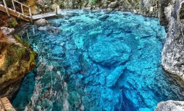 A crystal-clear, blue cenote with rocky walls and underwater stones—a highlight of tours en Punta Cana. A small wooden bridge extends over the water on the left, surrounded by rugged, mossy rock. The vivid blue water reveals rocks and depth, inviting tranquility.