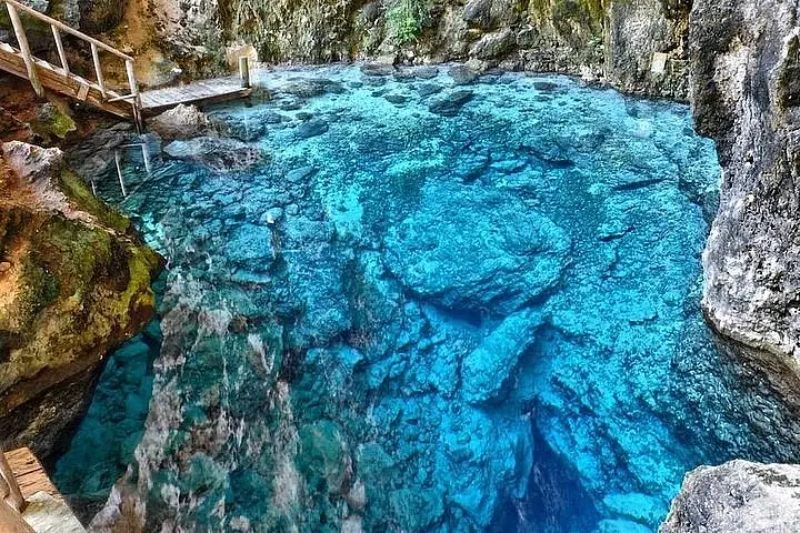A crystal-clear, blue cenote with rocky walls and underwater stones—a highlight of tours en Punta Cana. A small wooden bridge extends over the water on the left, surrounded by rugged, mossy rock. The vivid blue water reveals rocks and depth, inviting tranquility.