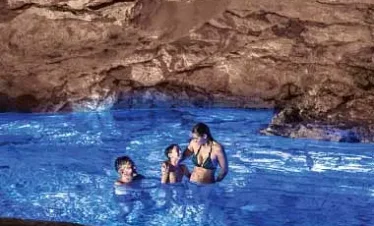 Three people stand together in clear, bright blue water inside a rocky cave. Sunlight from above creates shimmering reflections as they enjoy a swim—an unforgettable moment on one of many scenic Punta Cana excursions.