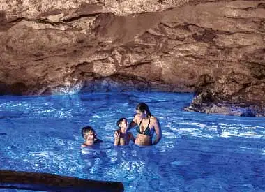 Three people stand together in clear, bright blue water inside a rocky cave. Sunlight from above creates shimmering reflections as they enjoy a swim—an unforgettable moment on one of many scenic Punta Cana excursions.