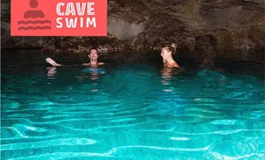 Two people swim in the bright turquoise water of an underground cave. The rocky ceiling is visible above them. A red sign in the top left reads “CAVE SWIM.” This hidden gem is a highlight of many popular tours in Punta Cana.