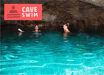 Two people swim in the bright turquoise water of an underground cave. The rocky ceiling is visible above them. A red sign in the top left reads “CAVE SWIM.” This hidden gem is a highlight of many popular tours in Punta Cana.