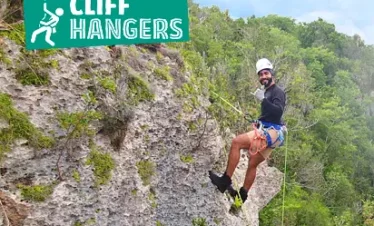 A smiling man wearing a helmet, harness, and climbing gear is rappelling down a rocky, moss-covered cliff face. He is surrounded by lush green trees—a perfect scene for adventurous Punta Cana excursions. A bright green “Cliff Hangers” sign is overhead.