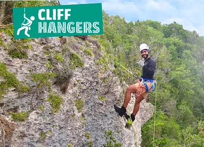 A smiling man wearing a helmet, harness, and climbing gear is rappelling down a rocky, moss-covered cliff face. He is surrounded by lush green trees—a perfect scene for adventurous Punta Cana excursions. A bright green “Cliff Hangers” sign is overhead.