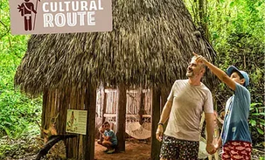 A man and a child hold hands and point towards the trees near a thatched hut in a lush, green forest. A “Cultural Route” sign hints at unique tours in Punta Cana, while hammocks and cultural artifacts fill the hut’s cozy interior.