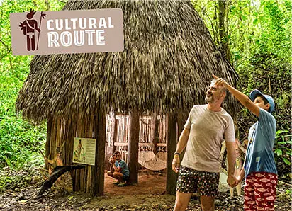 A man and a child hold hands and point towards the trees near a thatched hut in a lush, green forest. A “Cultural Route” sign hints at unique tours in Punta Cana, while hammocks and cultural artifacts fill the hut’s cozy interior.
