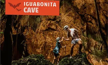 Two people, one adult and one child, wearing hats and casual clothes, climb mossy rocks inside a large cave with rugged brown walls. Above them, an orange “IGUABONITA CAVE” sign hints at adventure-filled tours in Punta Cana.