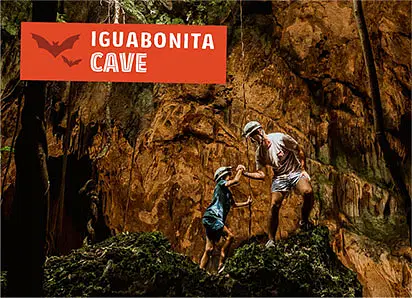 Two people, one adult and one child, wearing hats and casual clothes, climb mossy rocks inside a large cave with rugged brown walls. Above them, an orange “IGUABONITA CAVE” sign hints at adventure-filled tours in Punta Cana.