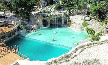 A natural turquoise pool surrounded by rocky cliffs and lush greenery, with small waterfalls flowing in. Wooden walkways border the pool, where a few people float peacefully—an idyllic spot often featured on Punta Cana excursions.