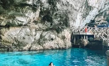 A person swims in clear turquoise water inside a large rocky cave. Stairs and a wooden walkway line the cave wall, where several people stand and observe—a stunning spot often explored on Punta Cana excursions. Sunlight brightens the vibrant blue water below.