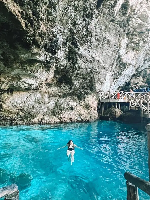 A person swims in clear turquoise water inside a large rocky cave. Stairs and a wooden walkway line the cave wall, where several people stand and observe—a stunning spot often explored on Punta Cana excursions. Sunlight brightens the vibrant blue water below.