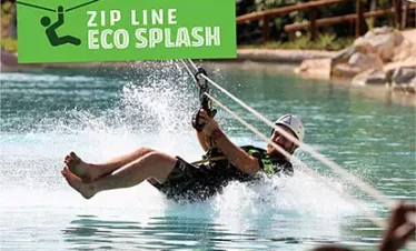 A person wearing a helmet rides a zip line over a pool, splashing water as they skim the surface. A green sign reads "ZIP LINE ECO SPLASH." Enjoy thrilling Punta Cana excursions surrounded by lush trees and rocks in the background.