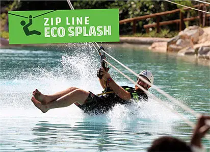 A person wearing a helmet rides a zip line over a pool, splashing water as they skim the surface. A green sign reads "ZIP LINE ECO SPLASH." Enjoy thrilling Punta Cana excursions surrounded by lush trees and rocks in the background.