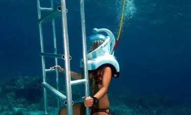 A person in a swimsuit and full-face snorkel mask climbs an underwater metal ladder during one of the popular Punta Cana excursions, holding the ladder with one hand while surrounded by clear blue water and vibrant coral below.
