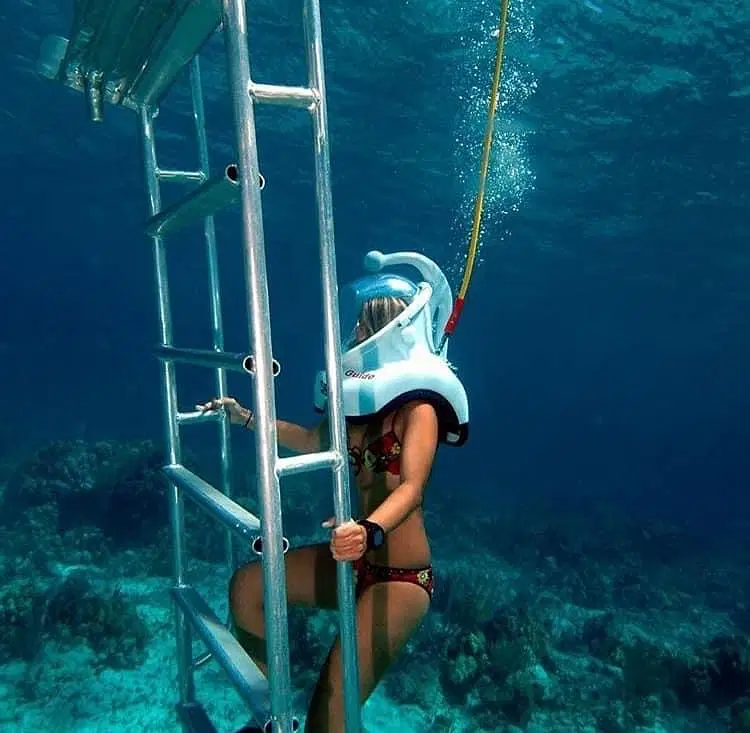 A person in a swimsuit and full-face snorkel mask climbs an underwater metal ladder during one of the popular Punta Cana excursions, holding the ladder with one hand while surrounded by clear blue water and vibrant coral below.
