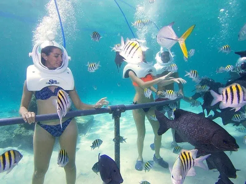 Two people underwater wear helmet-style diving gear and swimwear, standing behind a rail on one of the unforgettable Punta Cana excursions. They’re surrounded by colorful tropical fish in clear blue water, observing or interacting with them.