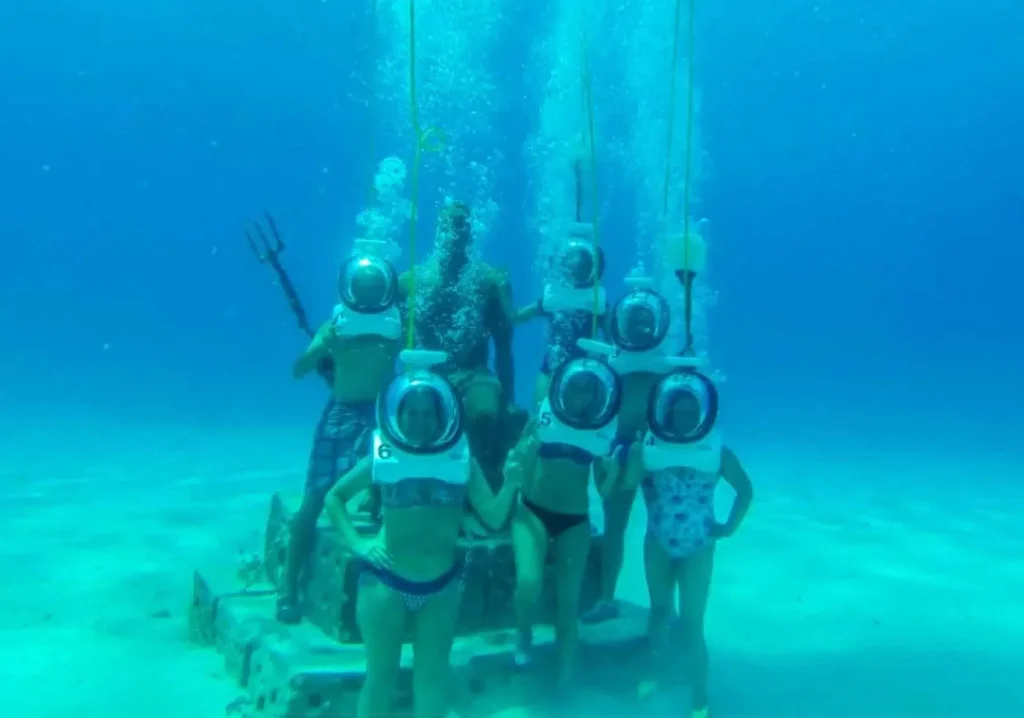 Six people in large, round diving helmets pose underwater around a seated statue holding a trident. Surrounded by the sandy seafloor and blue water, they enjoy one of the unique Punta Cana excursions—a memorable underwater tour full of adventure.