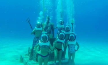 Six people in large, round diving helmets pose underwater around a seated statue holding a trident. Surrounded by the sandy seafloor and blue water, they enjoy one of the unique Punta Cana excursions—a memorable underwater tour full of adventure.