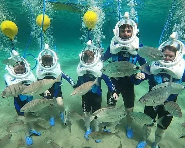 Five people in blue wetsuits and white diving helmets stand underwater during one of the exciting tours in Punta Cana, surrounded by swimming fish. They hold hands as air bubbles rise, with yellow buoys and blue hoses overhead connecting to their helmets.