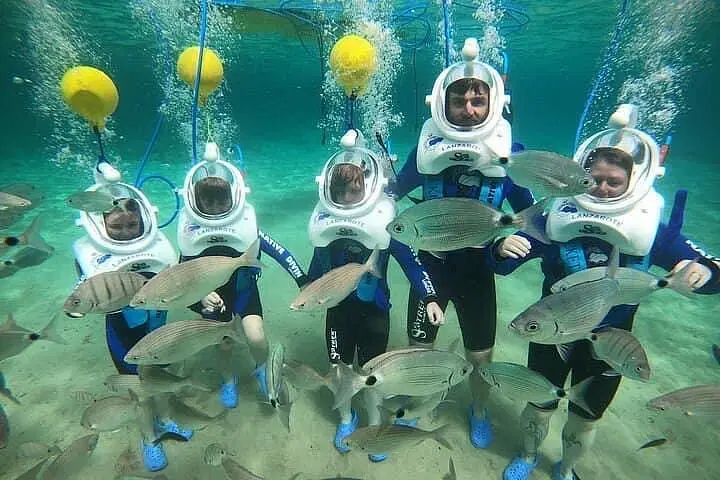 Five people in blue wetsuits and white diving helmets stand underwater during one of the exciting tours in Punta Cana, surrounded by swimming fish. They hold hands as air bubbles rise, with yellow buoys and blue hoses overhead connecting to their helmets.