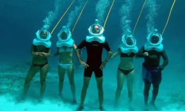 Five people stand underwater in swimsuits and white helmet diving gear, each connected by a yellow tube. Air bubbles rise as they pose on the sandy seafloor—an unforgettable experience offered on tours in Punta Cana.