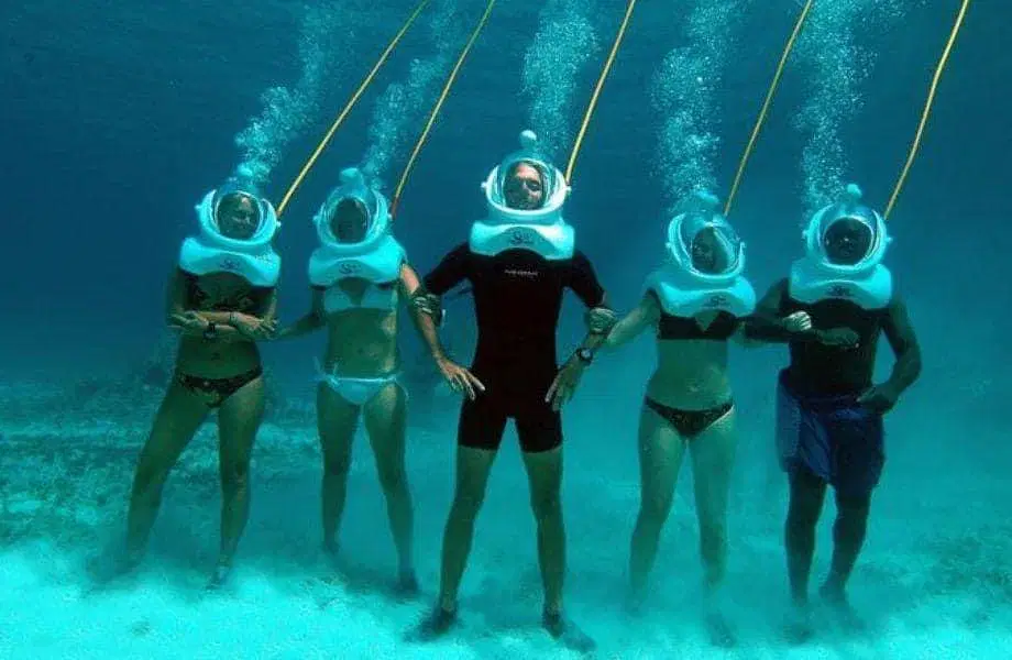 Five people stand underwater in swimsuits and white helmet diving gear, each connected by a yellow tube. Air bubbles rise as they pose on the sandy seafloor—an unforgettable experience offered on tours in Punta Cana.