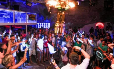 A crowded nightclub captures the vibrant Punta Cana nightlife, with people dancing and taking photos. Colorful lights illuminate a gold-costumed performer elevated in the center as partygoers in summer clothes enjoy drinks, creating a lively atmosphere.