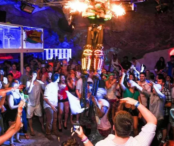 A crowded nightclub captures the vibrant Punta Cana nightlife, with people dancing and taking photos. Colorful lights illuminate a gold-costumed performer elevated in the center as partygoers in summer clothes enjoy drinks, creating a lively atmosphere.