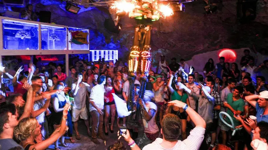 A crowded nightclub captures the vibrant Punta Cana nightlife, with people dancing and taking photos. Colorful lights illuminate a gold-costumed performer elevated in the center as partygoers in summer clothes enjoy drinks, creating a lively atmosphere.