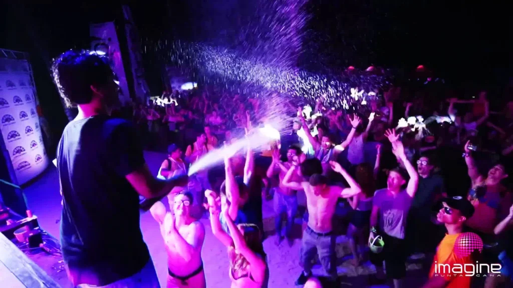 A crowd at a lively outdoor concert in Punta Cana dances and cheers as a performer sprays water over the audience. Colorful lights illuminate the scene, and many shirtless attendees revel in the energetic festival atmosphere.